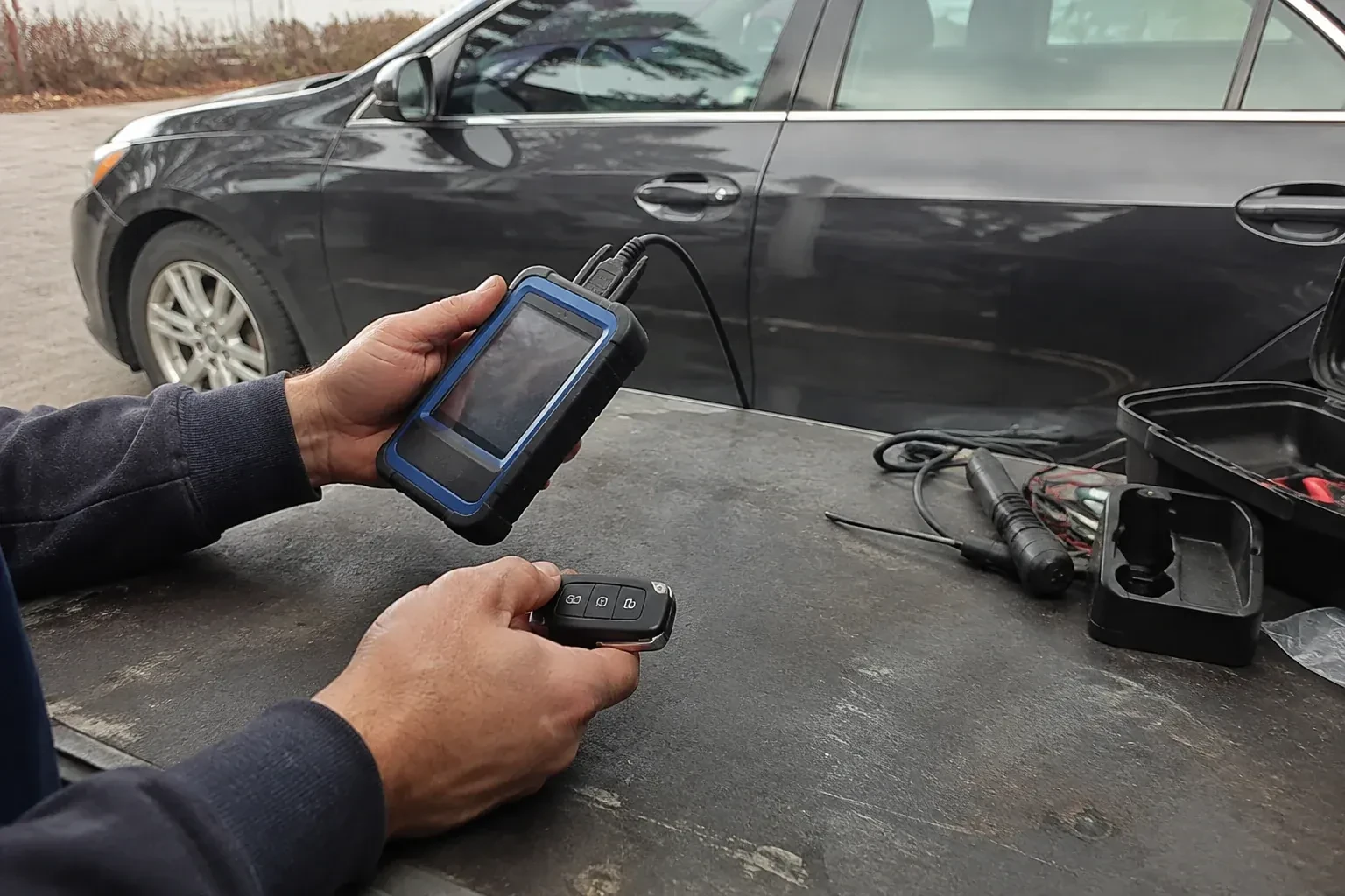 Hands holding a blue-edged electronic device and car-key fob at a worktable beside a parked car. — Transponder Key Programming, Oshawa