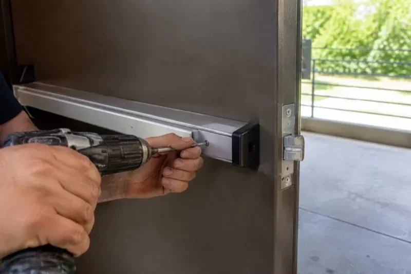 Hands using a power driver at the end of a horizontal push bar on an open metal door. — Panic & Exit Hardware, Oshawa